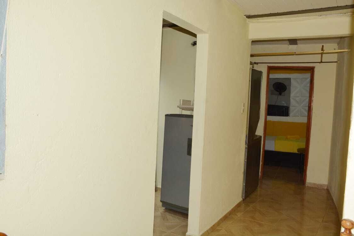 Un pasillo estrecho con suelo de baldosas, una puerta que da a una habitación con cama y ventana, y un refrigerador pequeño en la esquina.