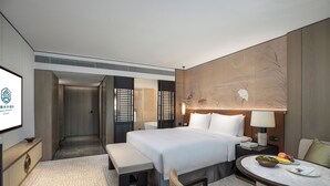 A modern hotel room with a large bed, bedside table, lamp, and a small round table with fruits and a bowl.