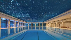 A large indoor swimming pool with a starry ceiling, large windows, and sun loungers along the sides.
