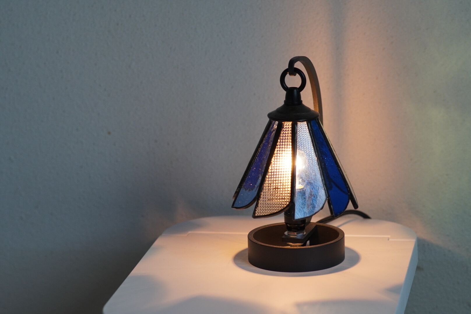 A table lamp with a blue glass shade and a black base, placed on a white surface against a plain wall.