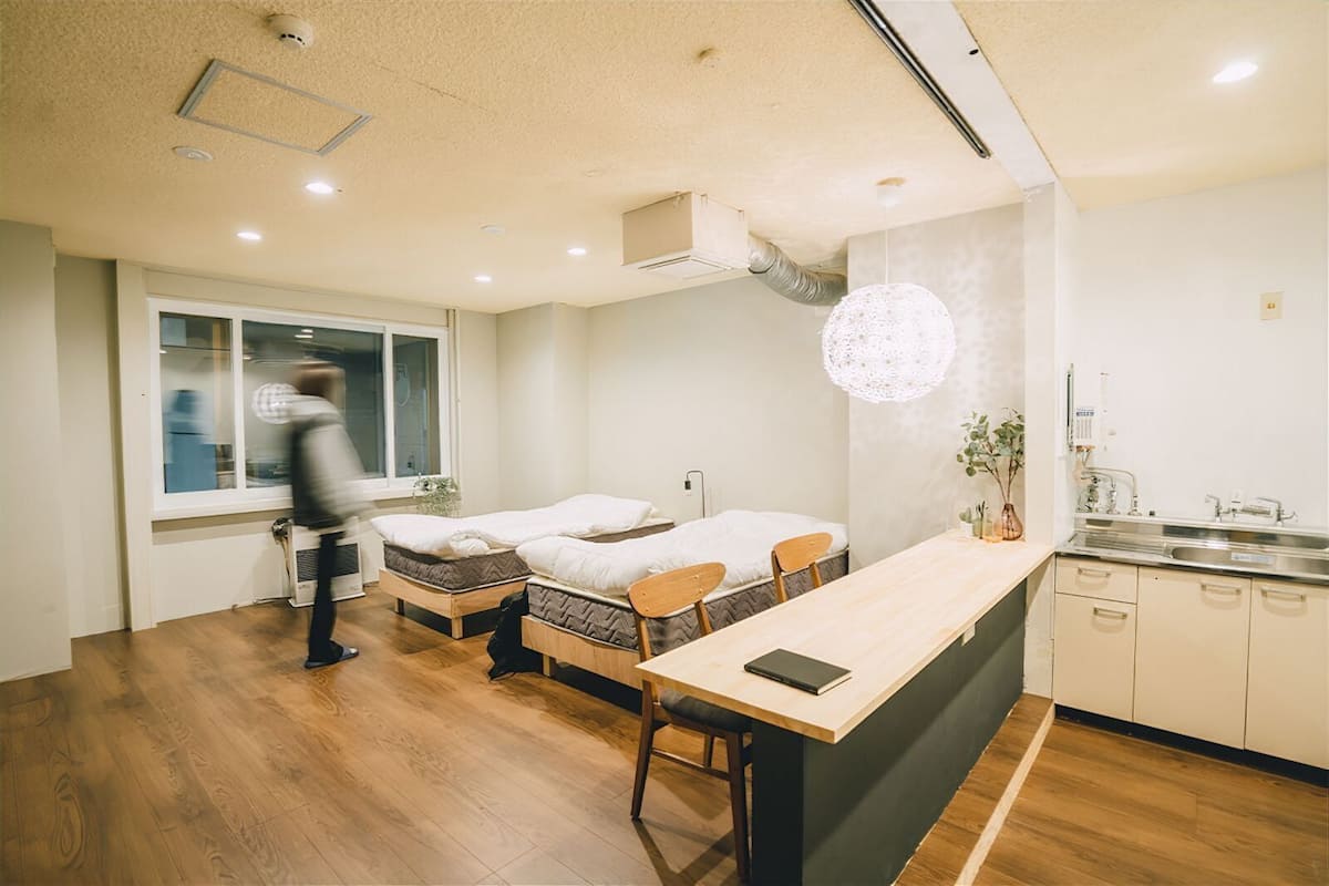 A modern room with two single beds, a wooden dining table, and a kitchen area with a sink and cabinets.