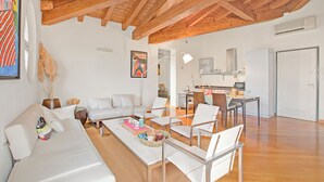 Un área de estar amplia con techo de madera, muebles blancos y una cocina al fondo.