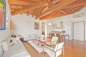 Uma sala de estar ampla com teto de madeira, móveis brancos e uma cozinha ao fundo.