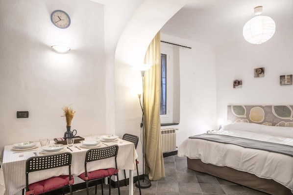 A room with a dining table set for two, a bed with a headboard, a clock on the wall, and a window with curtains.