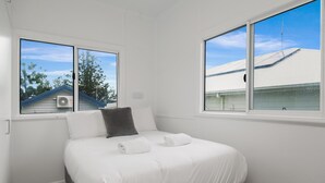 A bedroom with a bed, two windows, and a view of a house with a blue roof. - The Castaway (Emu Park)