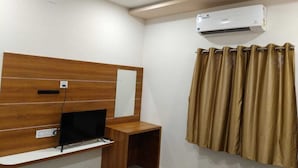 A hotel room with a wooden headboard, a flat-screen TV, a small wooden side table, an air conditioning unit, and a curtain. - Hotel Ayodhya Bhwan (Dwarka)