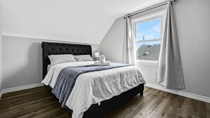 Ein Schlafzimmer mit dunklem Kopfteil, einem Bett mit weißer und grauer Bettwäsche, einem Fenster mit Vorhängen und Blick auf Gebäude.