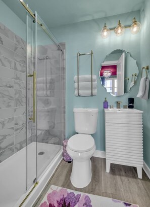 A bathroom with a glass shower enclosure, a white toilet, a sink with a mirror, and a light fixture above the mirror.