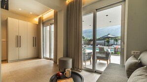 A modern hotel room with a sofa, a small table with a book and a mug, a sliding glass door leading to a balcony with a pool and outdoor furniture. - Villa Serenity- Modern villa for an exclusive vacation in Crete (Kournás)
