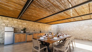 An outdoor kitchen with a stone wall backdrop, a wooden table set for dining, and a built-in refrigerator. - Villa Serenity- Modern villa for an exclusive vacation in Crete (Kournás)