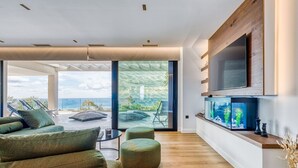 A modern living room with a large TV, green upholstered seating, and a view of the ocean.