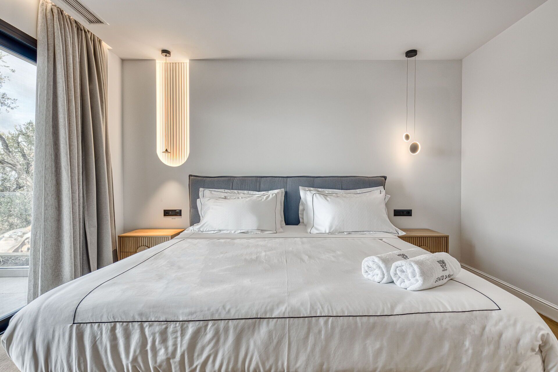 Une chambre d’hôtel moderne avec un grand lit, deux tables de chevet, une tête de lit et une fenêtre avec des rideaux.