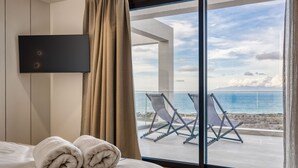 A hotel room with a bed, two rolled towels, a TV, and a balcony with beach chairs and a view of the sea.