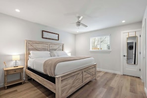 Une chambre à coucher avec un lit en bois, une table de chevet, un ventilateur de plafond, une fenêtre avec des stores et une porte dotée d’un miroir.