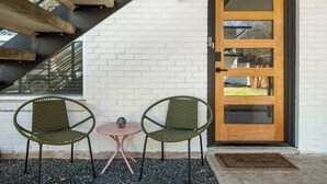 Un espacio al aire libre con dos sillas y una mesita, una puerta de madera con paneles de vidrio y un muro de ladrillo blanco.