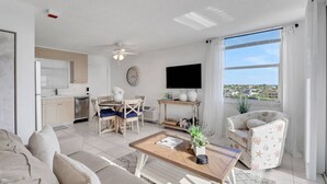 A modern living room with a sofa, coffee table, and chairs, a kitchen area with a dishwasher, and a large window with a view of a residential area.