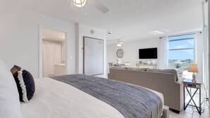 A modern hotel room with a large bed, a seating area with a sofa and table, a flat-screen TV, and a view of the outdoors.