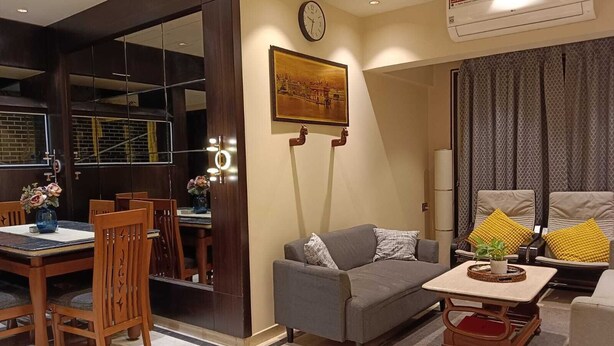 A modern living room with a sofa, a dining table with chairs, a clock, and an air conditioning unit. - Luxurious Sea-view 3 BHK Service Apartment In CBD Belapur. Roy Bari, Satya Stays (Navi Mumbai)