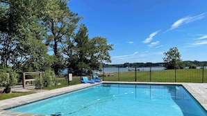 Une piscine rectangulaire avec une terrasse en pierre, entourée d’une clôture en métal noir, et offrant une vue sur un champ verdoyant et des arbres en arrière-plan.