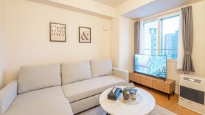 En moderne stue med en beige sofa, et sofabord med pyngeting, et fladskærms-TV og et stort vindue med gardiner.