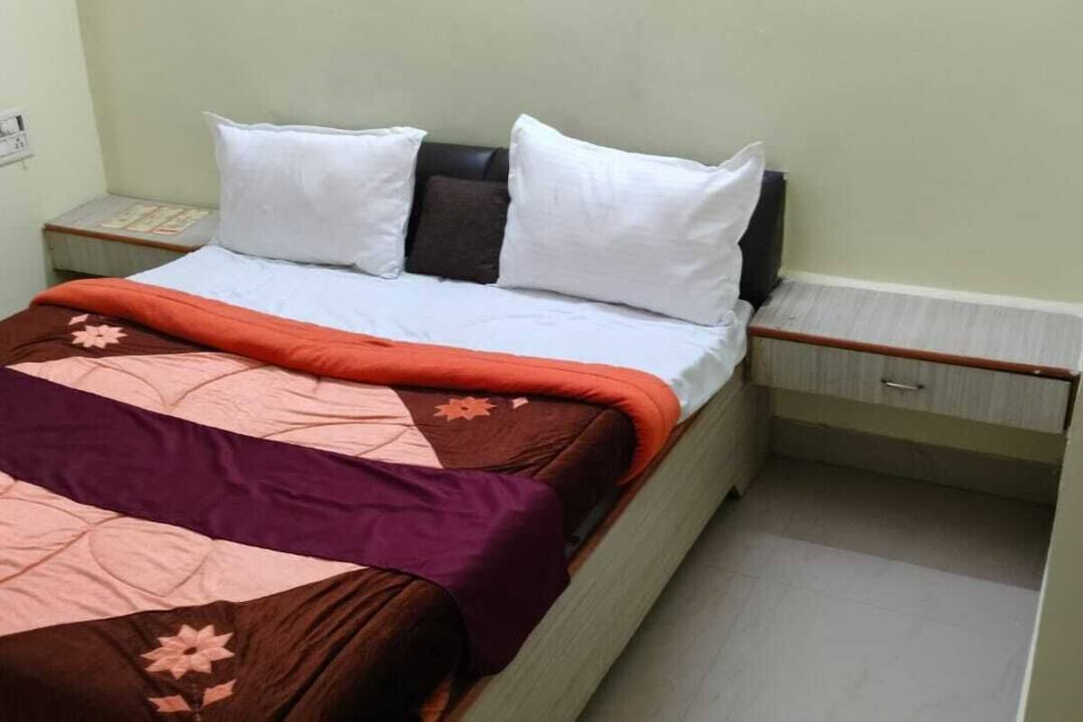 A single bed with white and dark pillows, a brown and orange bedspread, and a nightstand with a lamp.