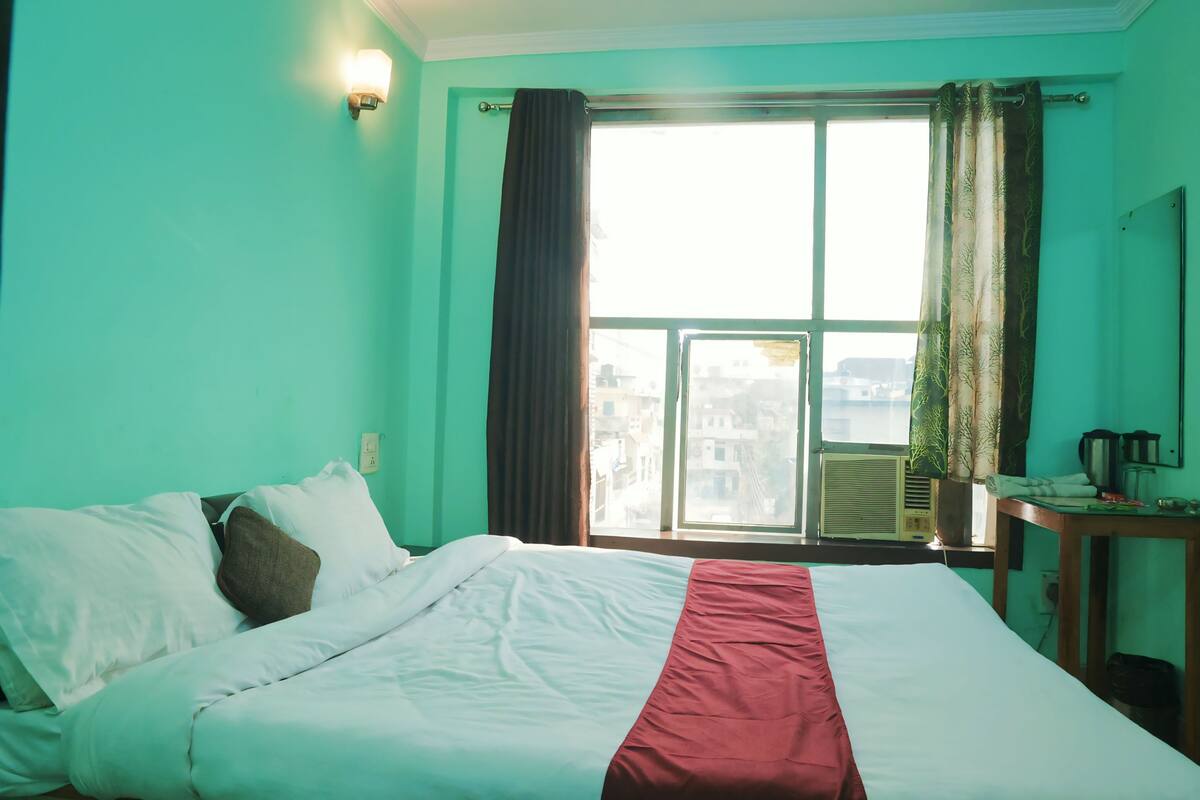 A hotel room with a bed, a window with curtains, a wall-mounted light, and an air conditioning unit.