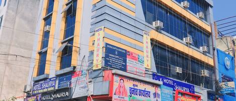 Un edificio de varios pisos con letreros que indican 'Hotel Shaurya' y 'K. J. Hotel Shaurya'.