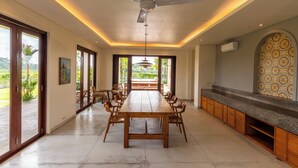 A spacious dining area with a long wooden table and chairs, a ceiling fan, and a view of the outdoors through large sliding doors. - Atelier Home by Great Bali Villas (Selemadeg)