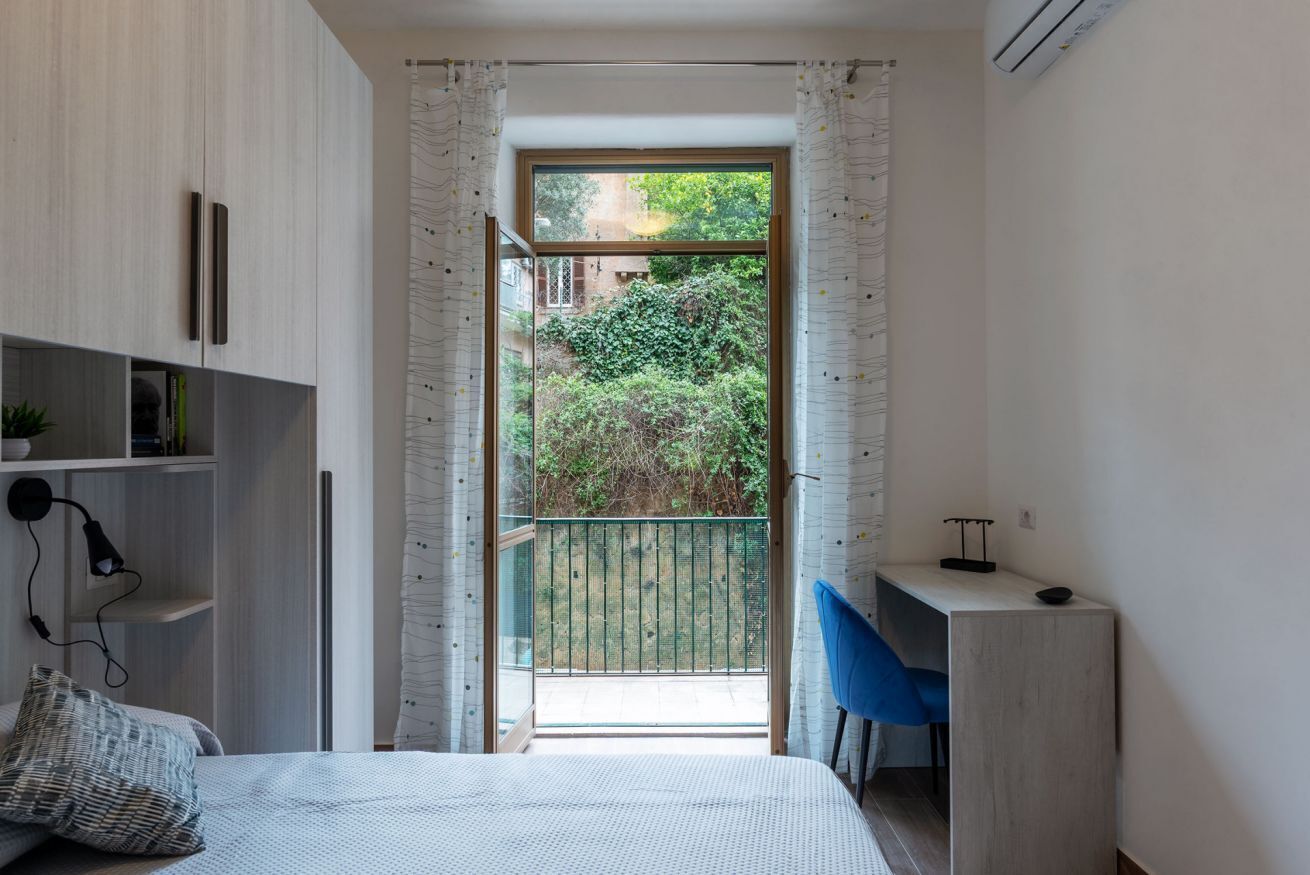 A bedroom with a bed, a desk, and a view of a balcony with greenery.