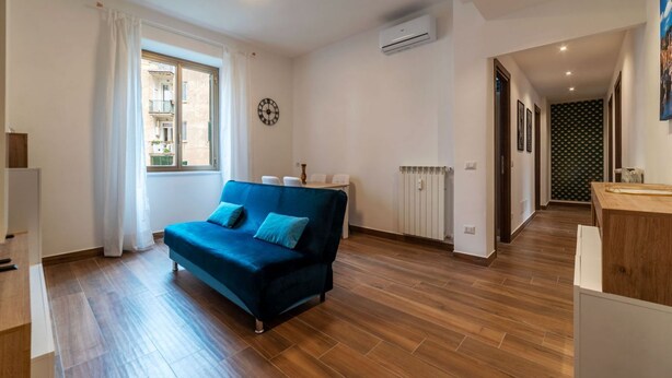 A living room with a blue sofa, wooden flooring, a window with curtains, an air conditioning unit, a clock, and a hallway leading to another room. - GuestHost - Garbatella Apartment - Roma (Roma)