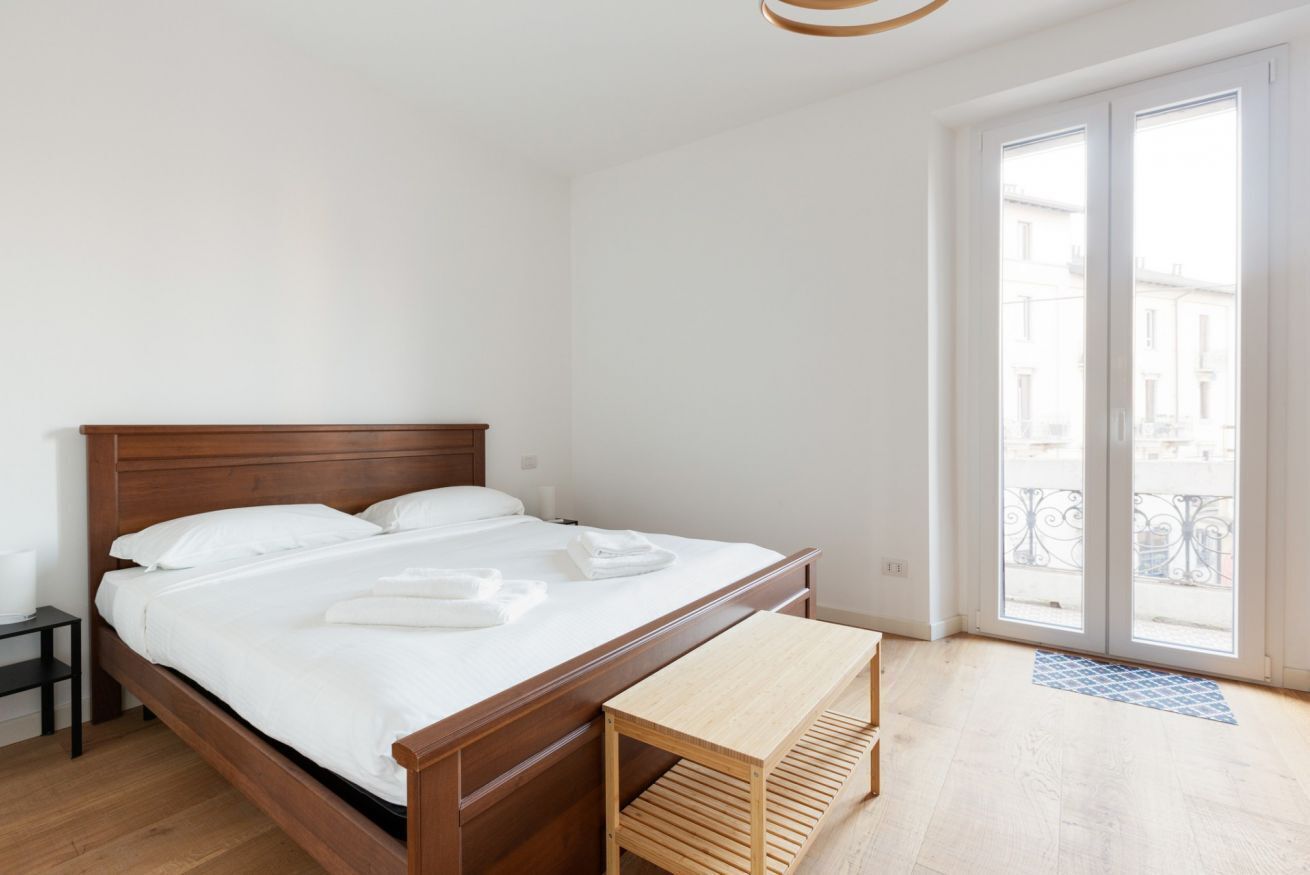 Ein Schlafzimmer mit einem hölzernen Bett, einem Nachttisch, einer Bank und Blick durch eine Glastür nach draußen.