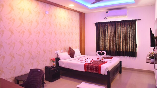 A hotel room with a bed, a desk, a chair, a ceiling fan, and a window with curtains. - RD Hotel (Perundurai)
