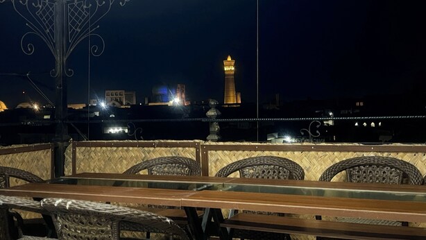 An outdoor seating area with wicker chairs and a wooden table, overlooking a cityscape at night with a prominent tower in the background. - SHOM EAST HOTEL (Bukhara)