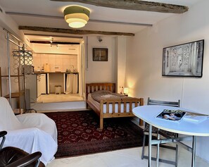 Un dormitorio con una cama, una mesita con una revista, una silla, un tapete rojo estampado y un pasillo que lleva a una cocina.
