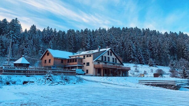 A large, two-story wooden house with a sloped roof, surrounded by a snow-covered landscape and dense forest.