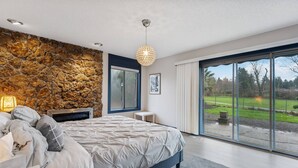 A modern bedroom with a stone wall, a large bed, a small table, and a view of a green landscape through large windows. - Incredible Golfside Single Level Ranch, Open Layout w/ Vintage Charm, Huge Patio, Close 2 Everything (Gresham)