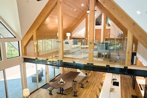 Um amplo loft com teto alto, vigas de madeira e guarda-corpo de vidro com vista para uma área de refeições.