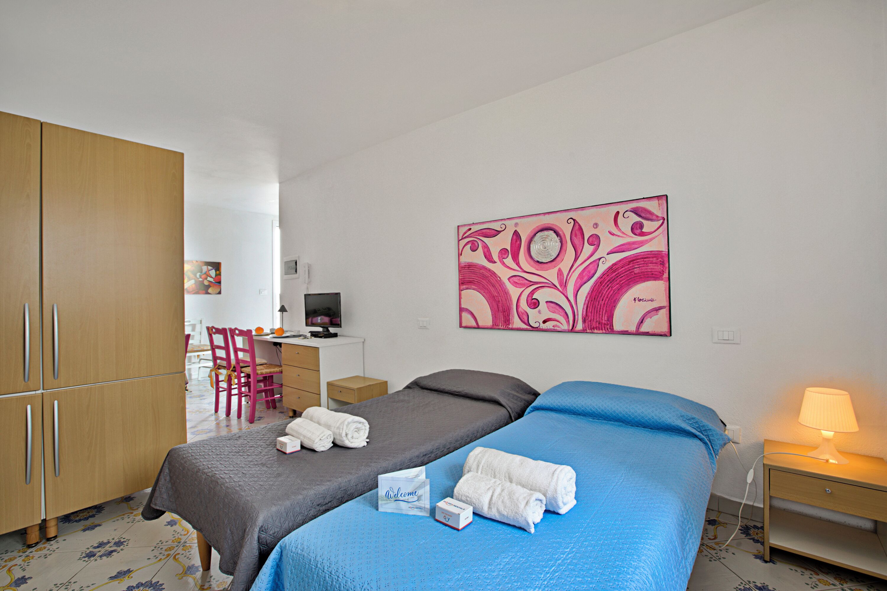 Een hotelkamer met twee bedden, een bureau, een stoel, een kledingkast en een schilderij aan de muur.