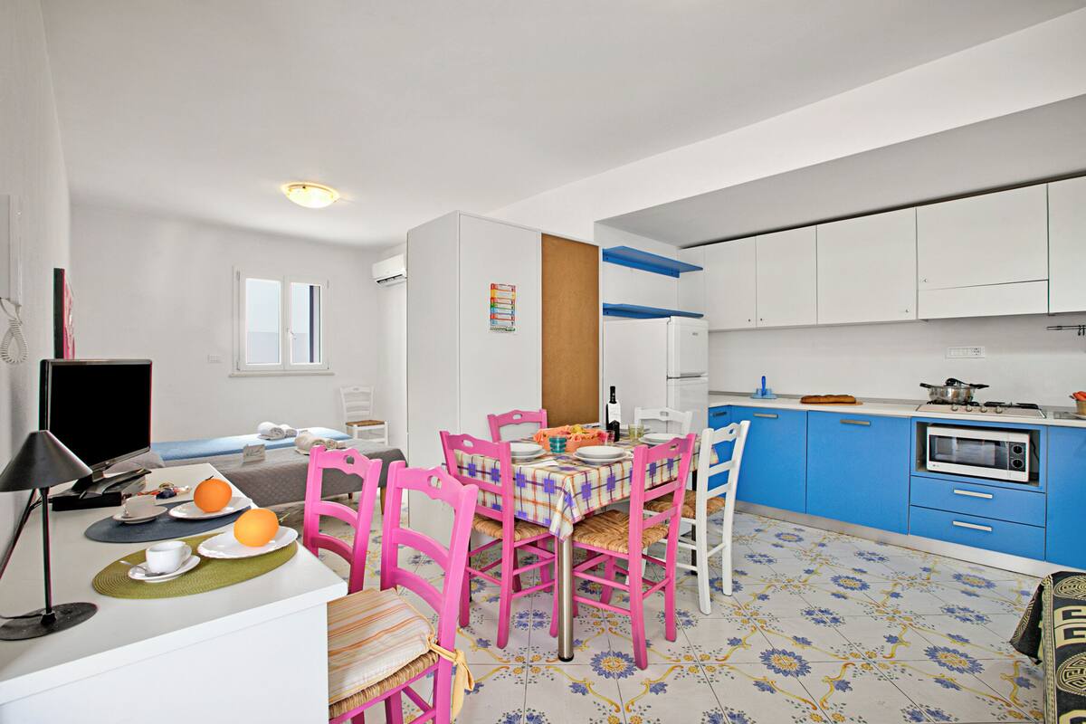 A compact kitchen with white cabinets, a blue countertop, and a dining area with pink chairs and a table set for two.
