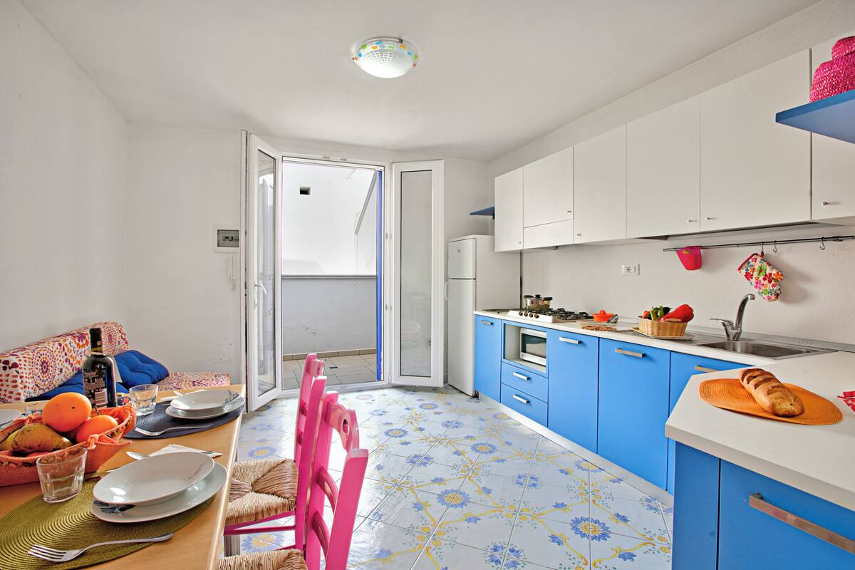 A compact kitchen with blue cabinets, a dining area with a pink chair, and a small table set with plates and a basket of fruit.