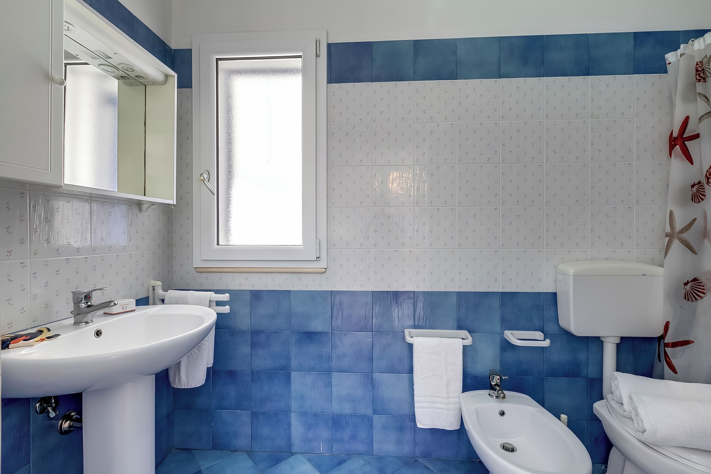 A bathroom with a sink, bidet, and toilet, featuring blue and white tiles and a window.