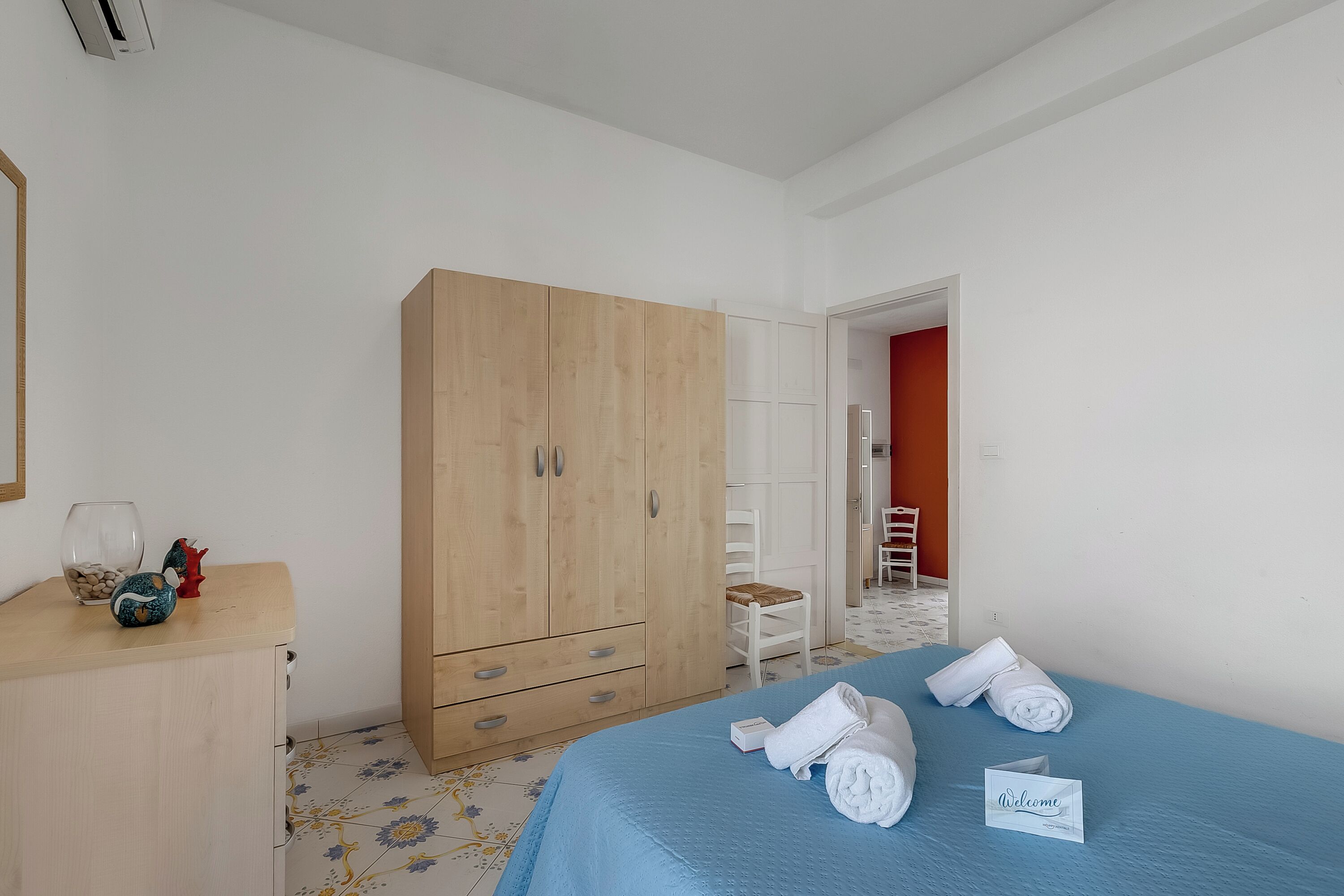 A bedroom with a bed, a wooden wardrobe, a small table with decorative items, and a chair.