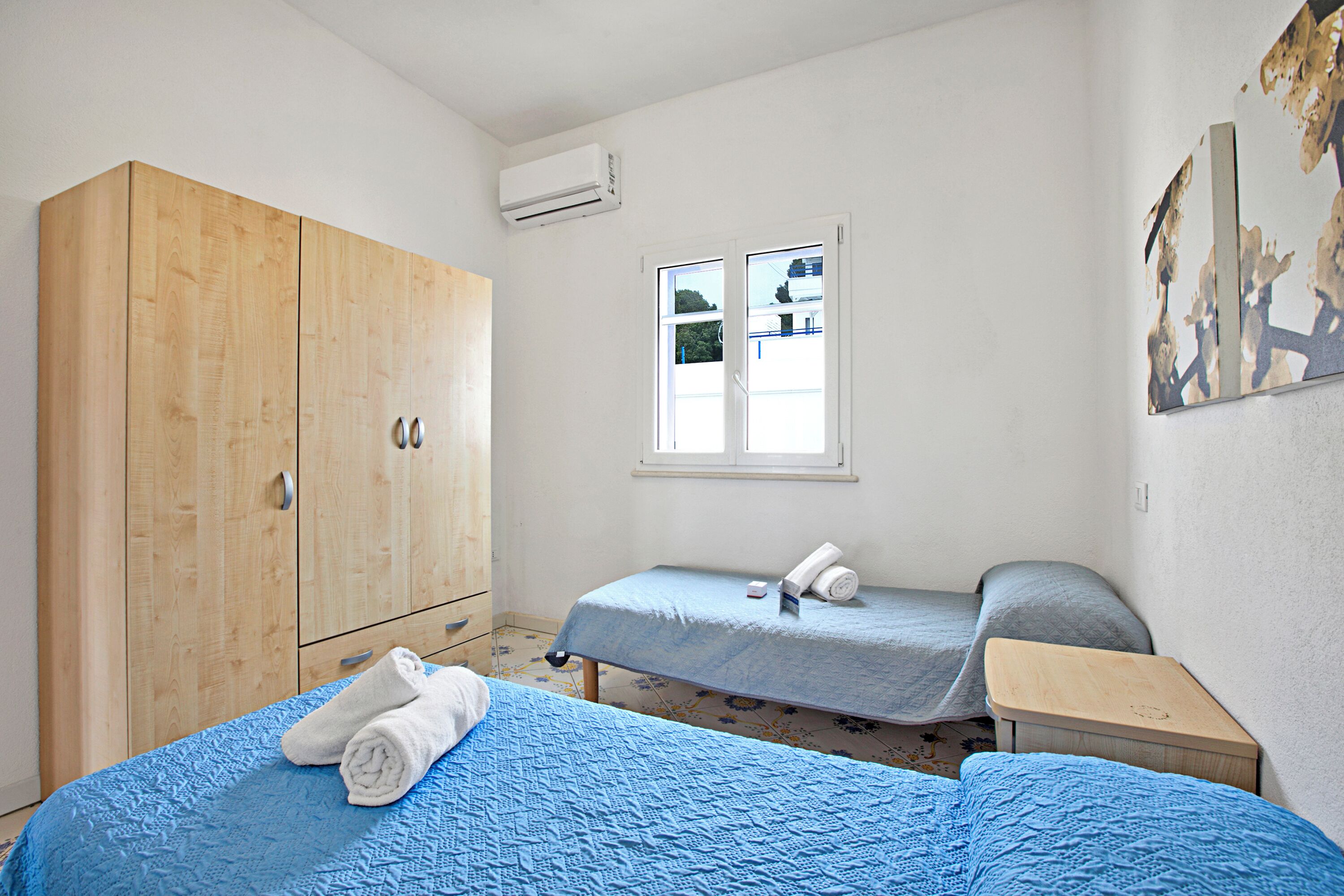 A bedroom with a wooden wardrobe, a single bed with a blue quilt, a bedside table, and a window with a view of the outside.