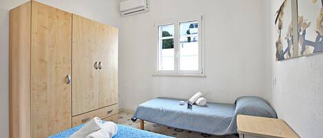 A bedroom with a wooden wardrobe, a single bed with a blue quilt, a bedside table, and a window with a view of the outside.