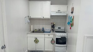 A compact kitchen with white cabinets, a sink, a stove, and an oven. There are kitchen utensils hanging on the wall and a doormat with a chef's hat on the floor.