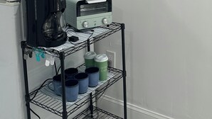A kitchenette with a white refrigerator, a coffee maker, a toaster, and a shelf with mugs and containers.
