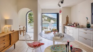 A modern kitchen with a view of the sea, a dining area with a round table, and a cozy seating area with a red cushion. - MARLI Exceptional Sea-View Villa with Private Pool – Authentic Cap Corse (Brando)