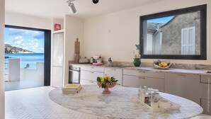 A modern kitchen with a marble countertop, a large window with a view of the sea, and a dining area with a table set for two. - MARLI Exceptional Sea-View Villa with Private Pool – Authentic Cap Corse (Brando)