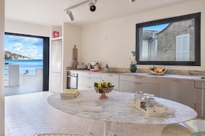 A modern kitchen with a marble countertop, a large window with a view of the sea, and a dining area with a table set for two.
