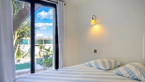 A bedroom with a bed, striped pillows, a nightstand, a wall-mounted lamp, and a view of a pool and greenery outside. - MARLI Exceptional Sea-View Villa with Private Pool – Authentic Cap Corse (Brando)